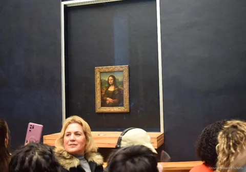 Salonun koyu duvarında cam arkasındaki Mona Lisa önünde ziyaretçiler, Louvre Müzesi biletleri ile turun öne çıkan anı