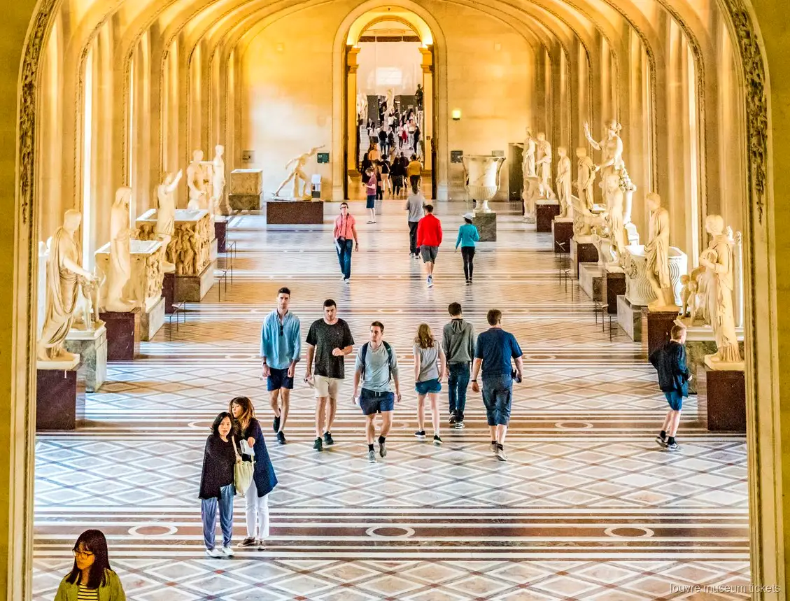 Visitantes en una galería de esculturas clásicas con arcos y suelo de mármol, vista con entradas Museo del Louvre